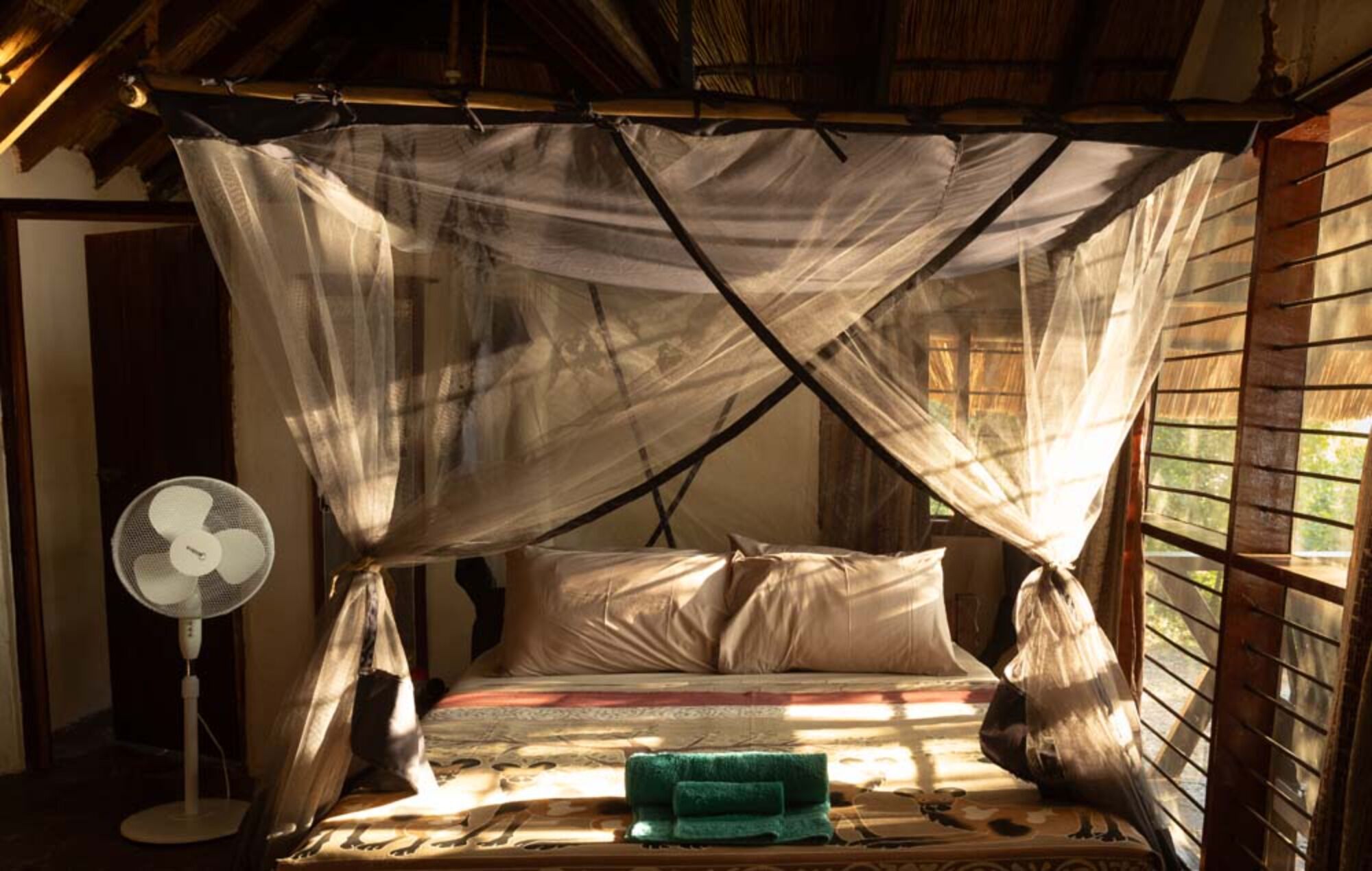 Una habitación con una cama bajo una mosquitera, un ventilador de pie y una ventana con contraventanas de madera.