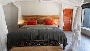 A bedroom with a canopy bed, a wooden headboard, bedside lamps, and a view of the outside. - Zikomo Safari (South Luangwa National Park)