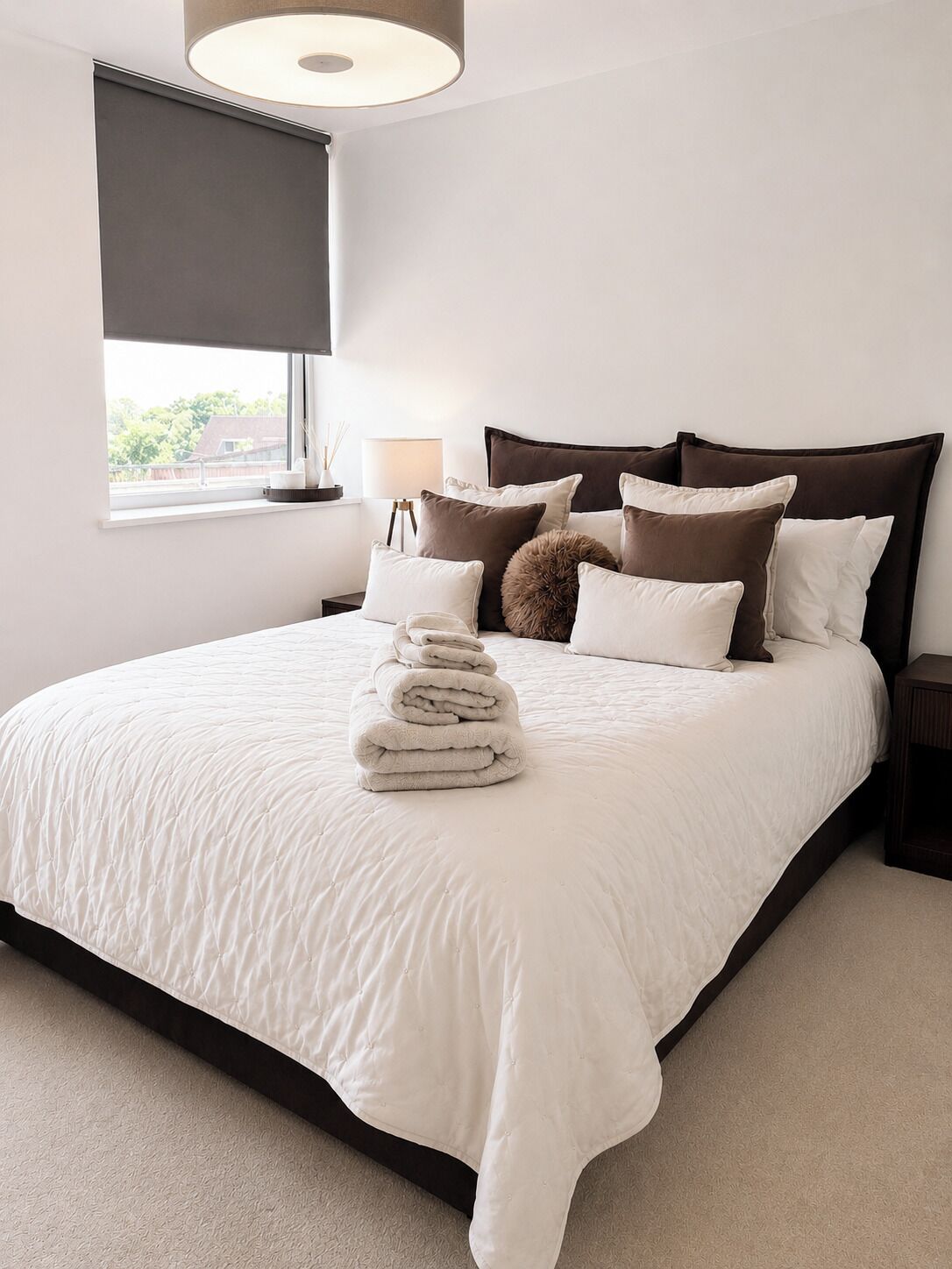A neatly made bed with a white duvet, a stack of folded towels, and a variety of pillows. A window with a blind is visible, and a nightstand with a lamp is placed beside the bed.