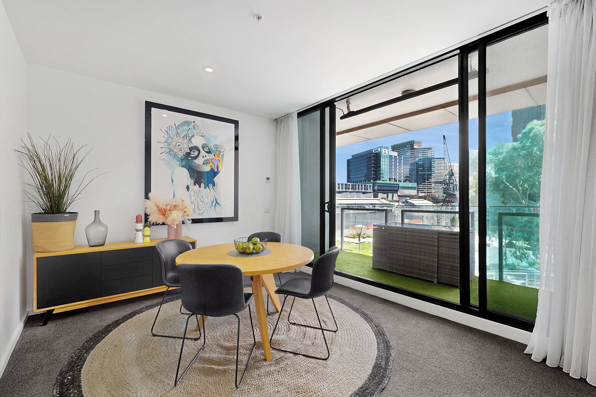 A modern dining area with a round table, black chairs, a potted plant, a vase, and a framed artwork on the wall.
