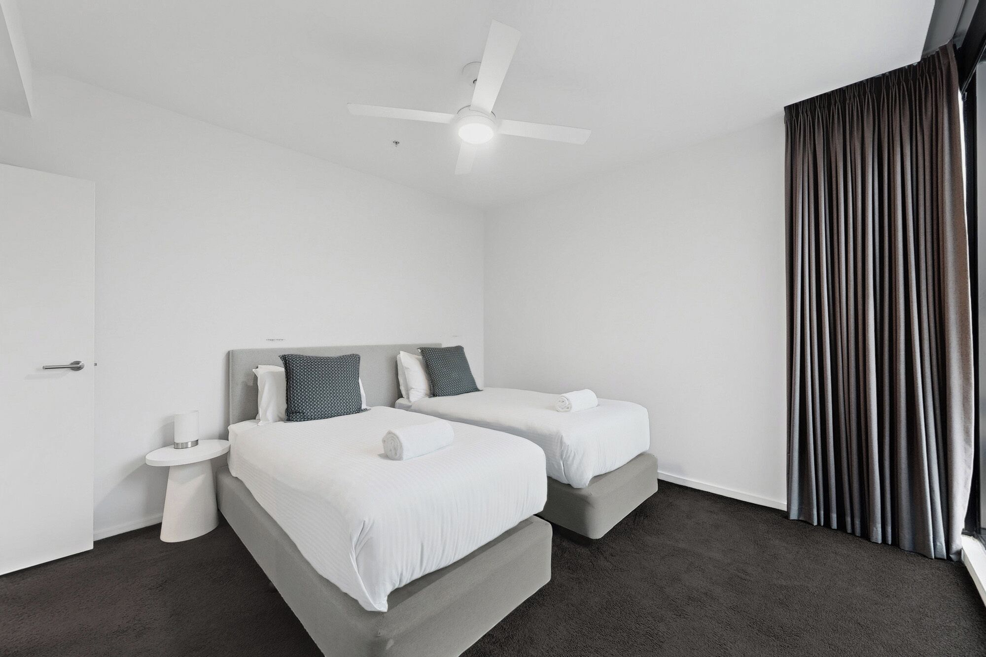 A hotel room with two beds, a ceiling fan, a window with curtains, and a white door.