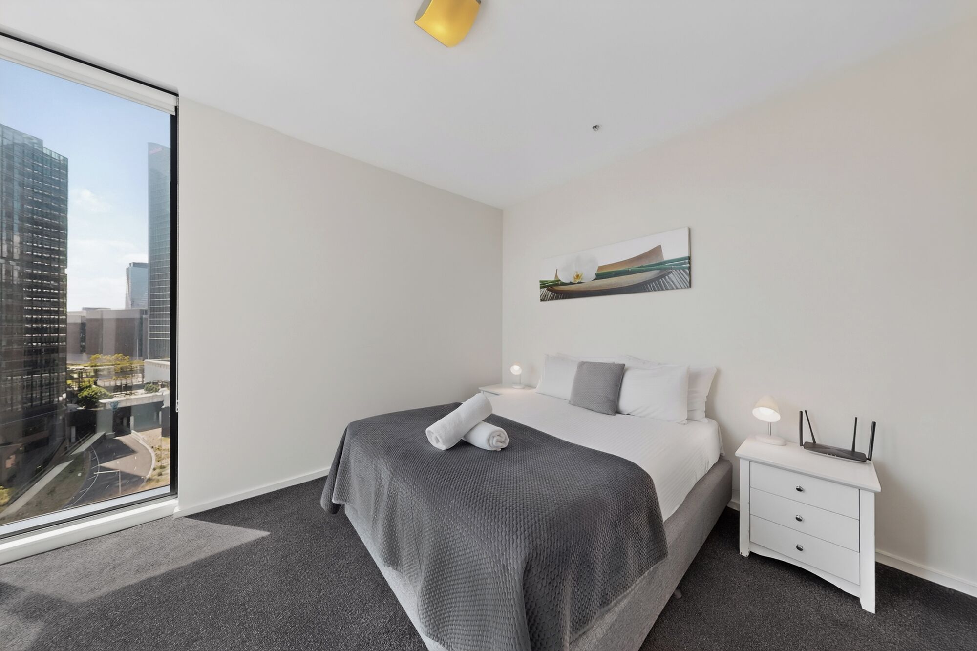 Una habitación de hotel moderna con un ventanal grande, una cama con un edredón gris con textura, una mesita de noche blanca con lámpara y un cuadro en la pared.