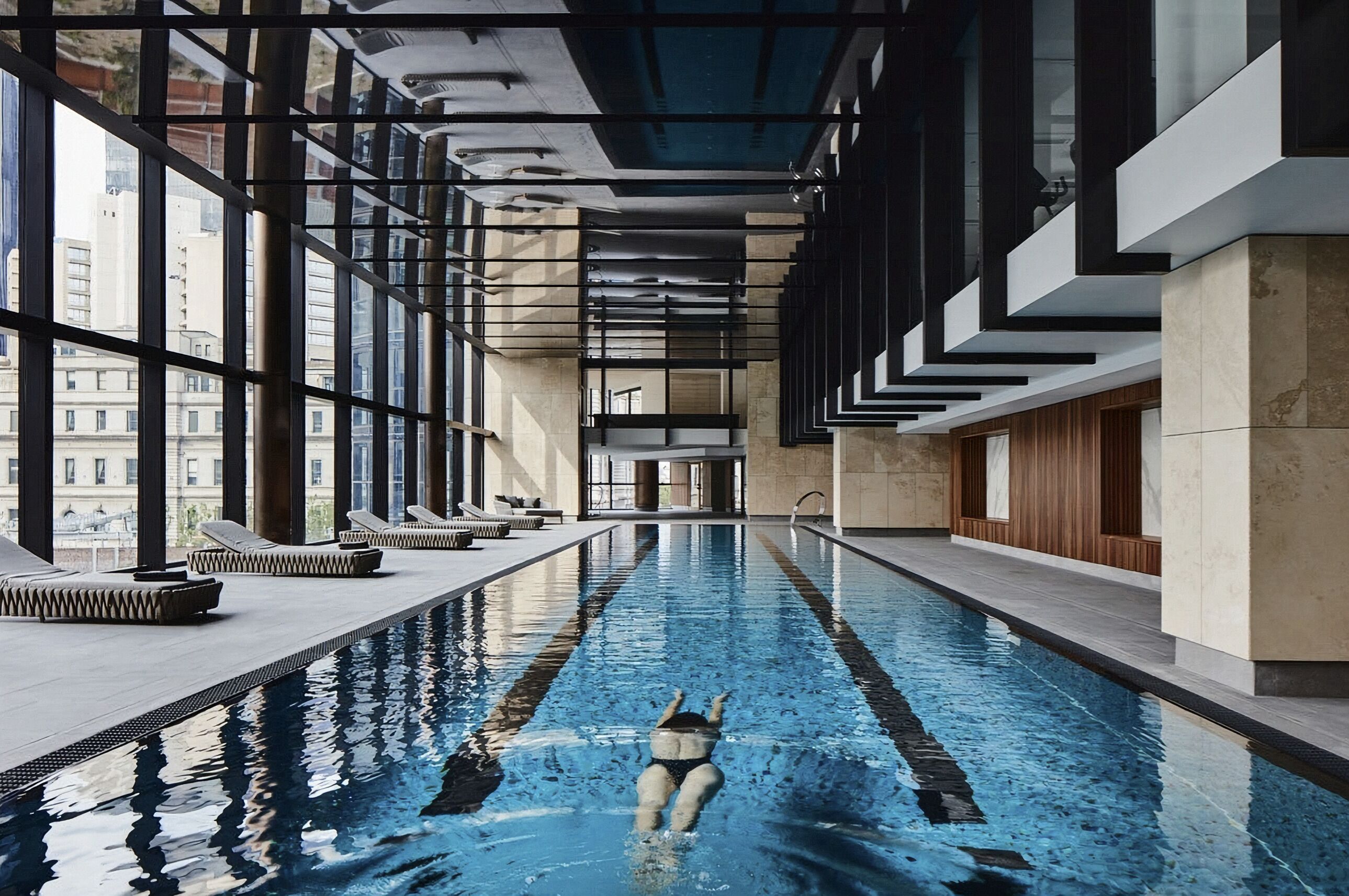 An indoor swimming pool with clear lanes, a person swimming, and a view of the city through large windows.