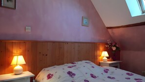 Een slaapkamer met een schuine balkenlaag, houten lambrisering, een bed met een bloemenbedekking, twee bedlampen en een vaas met bloemen.