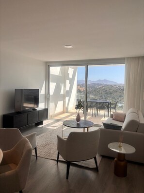 Ein modernes Wohnzimmer mit einer großen Schiebeglastür, einem Flachbildfernseher, einer Couch, einem Couchtisch und einem Essbereich mit Außenmöbeln.