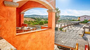 Eine Terrasse mit Tisch und Stühlen, Blick auf die Landschaft und ein Gebäude mit orangenem Mauerwerk.
