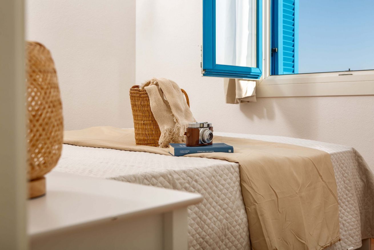 Ein Zimmer mit einem Bett, einer Kamera auf einem Buch, einem Korbstuhl und einem Fenster mit blauen Rollläden.
