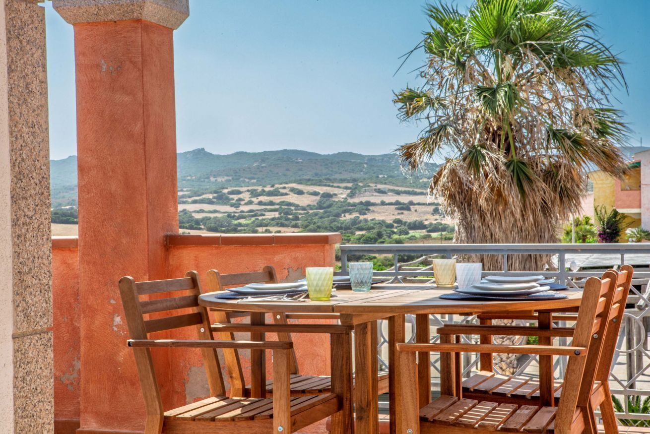 A wooden table and chairs on a balcony with a view of a palm tree and distant hills.
