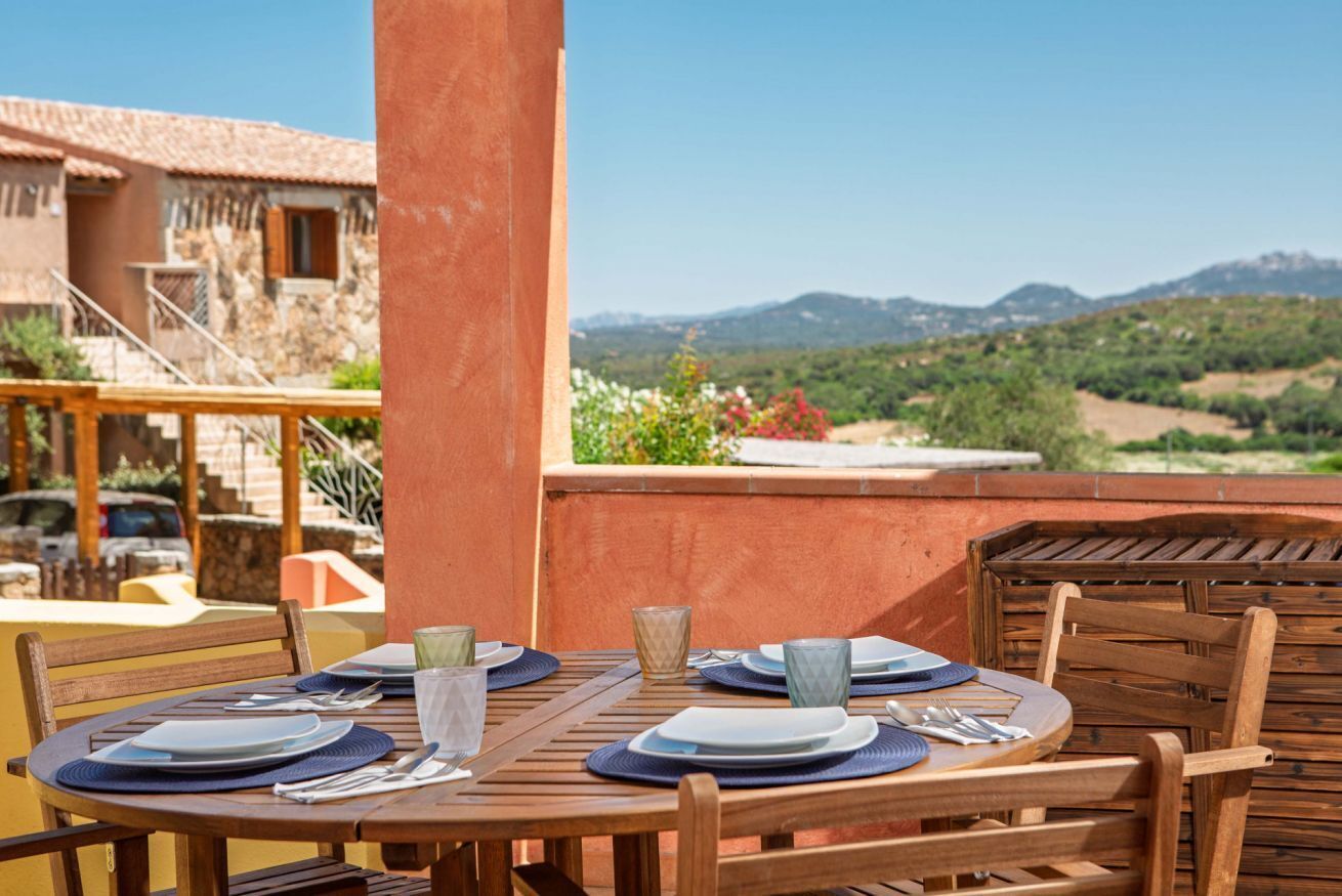 Una mesa de madera dispuesta para comer, con platos, cubiertos y copas, con vistas a un paisaje montañoso.