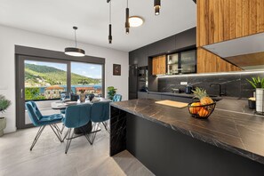 A modern kitchen with a marble countertop, a dining area with blue chairs, and a view of a hillside. - Alba Azzura 2-bedroom apartment in Zaton (Zaton)
