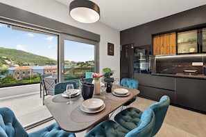 A modern dining area with a view of a lake and hills, a kitchen with dark cabinetry, and a dining table set for four. - Alba Azzura 2-bedroom apartment in Zaton (Zaton)