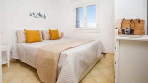 Een slaapkamer met een bed, een raam, een plank met een camera en een tas, en een wanddecoratie.