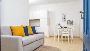 Een knus woongedeelte met een bank, een eettafel met stoelen, een keukenhoek en een blauwe deur.