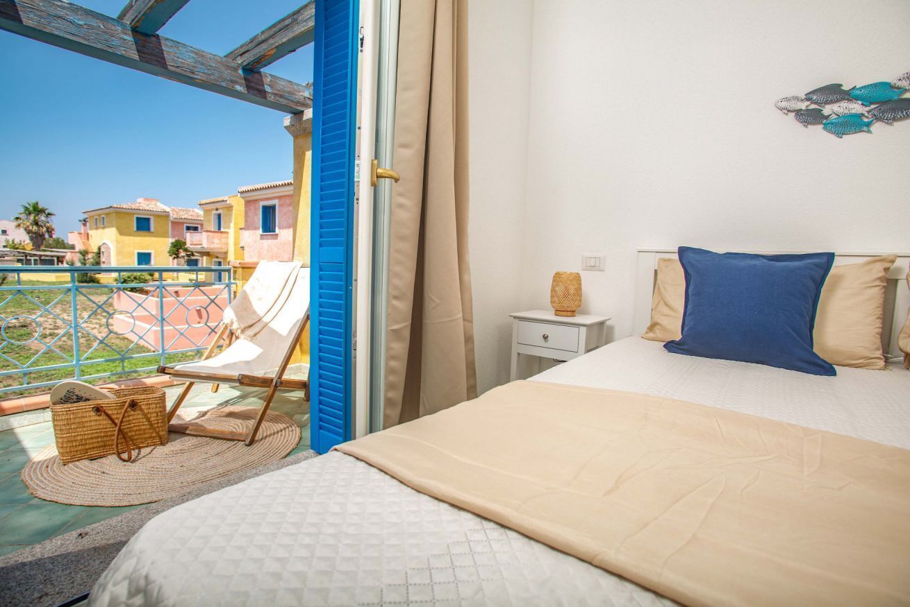 Une chambre à coucher comprenant un lit, une table de chevet, une chaise, un panier en osier et une vue sur les bâtiments depuis un balcon.
