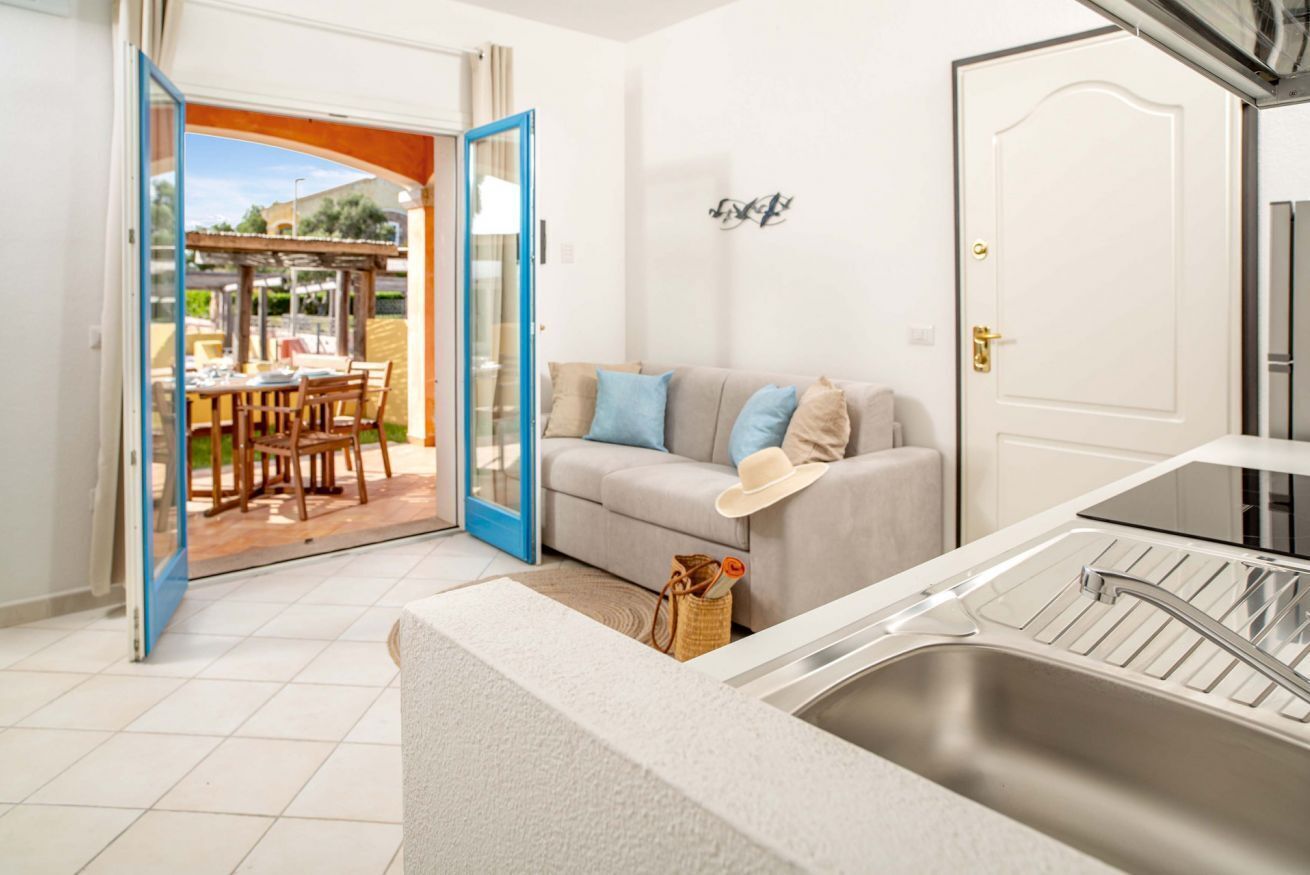 An open door leading to a patio with a table and chairs, a beige sofa with cushions, a white door, and a kitchen sink.