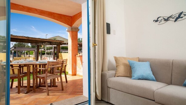Una habitació amb un sofà beix, una taula de dinar amb cadires i una porta oberta que dona a un patí exterior amb una pérgola.