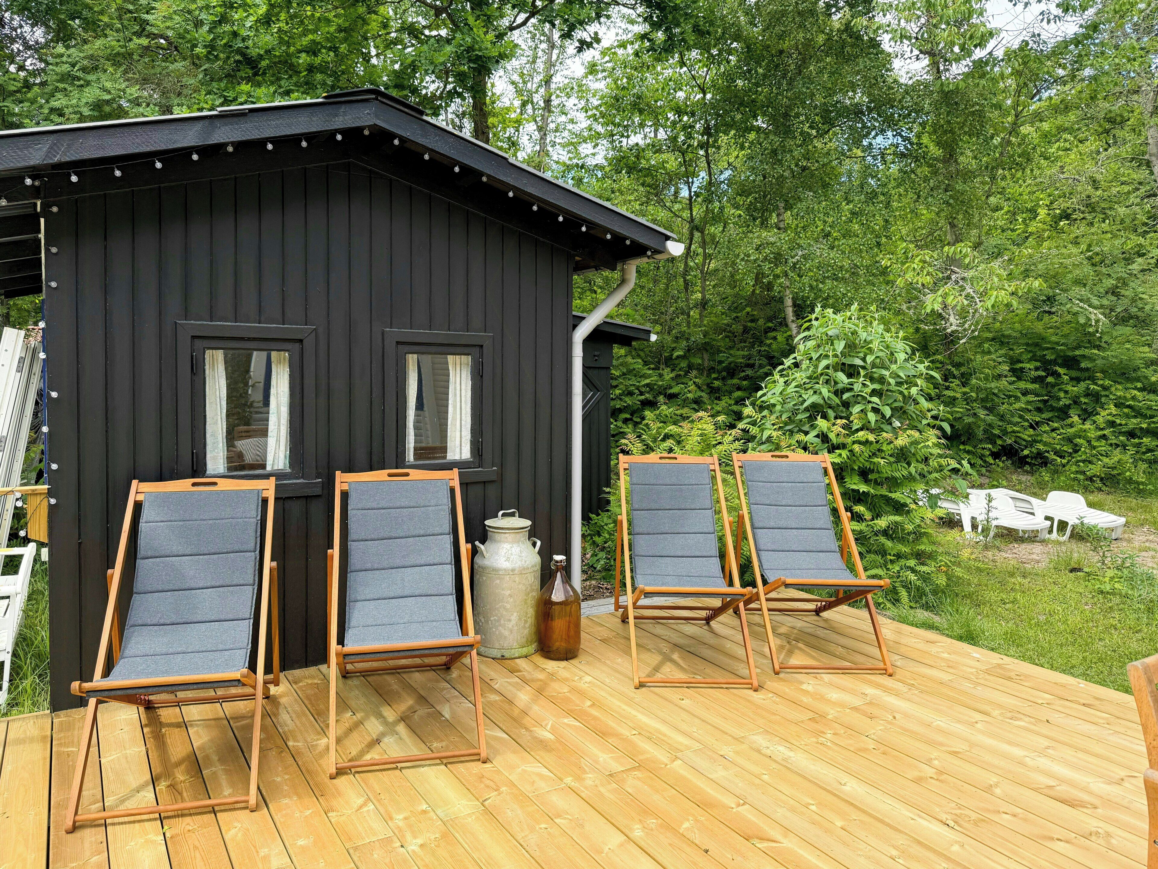 A wooden deck with four lounge chairs, a small black building with windows, and a lush green backdrop.
