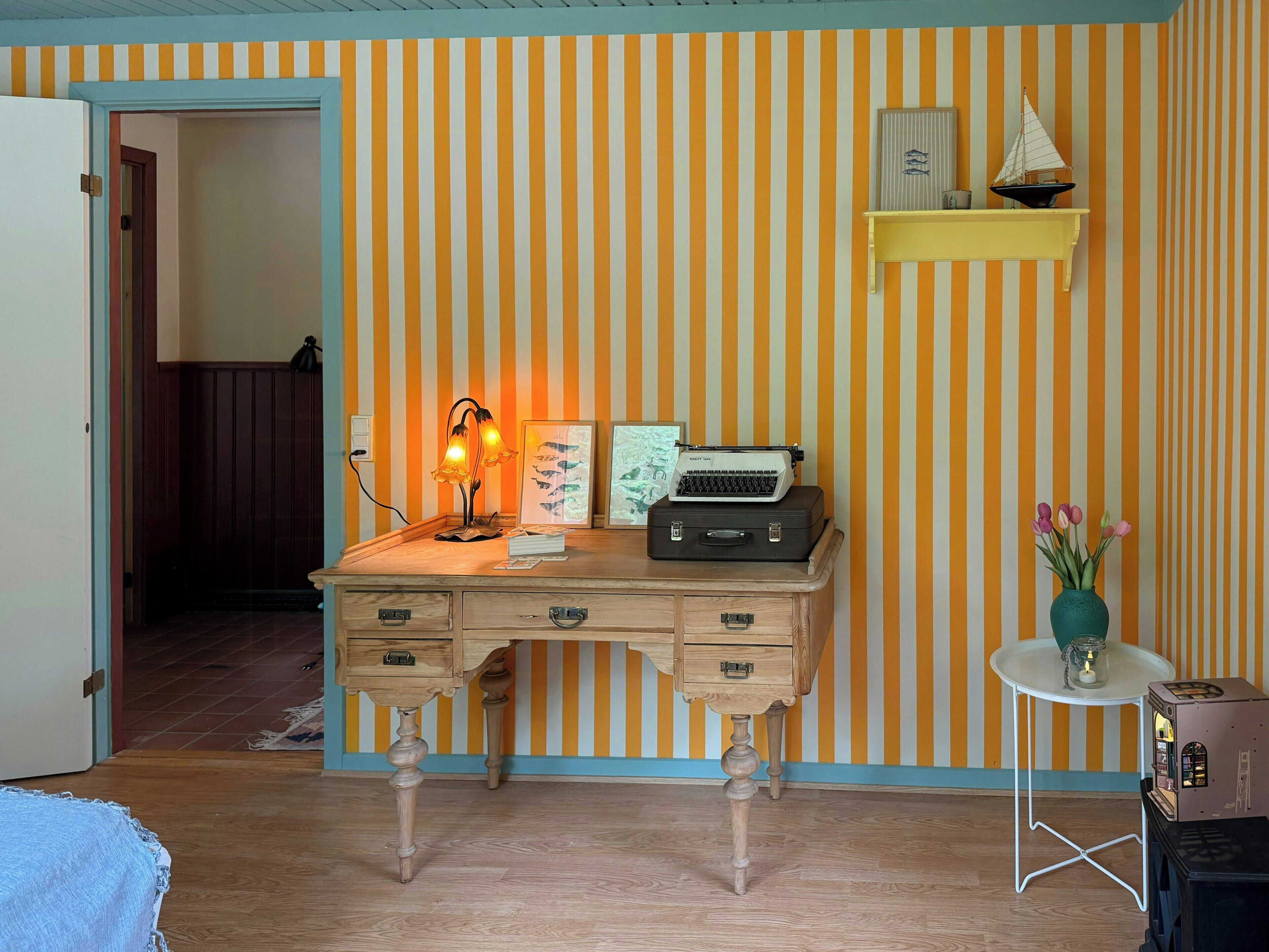 A room with a striped wallpaper, a desk with drawers, a typewriter, a lamp, a small shelf with a model ship, and a vase with flowers.