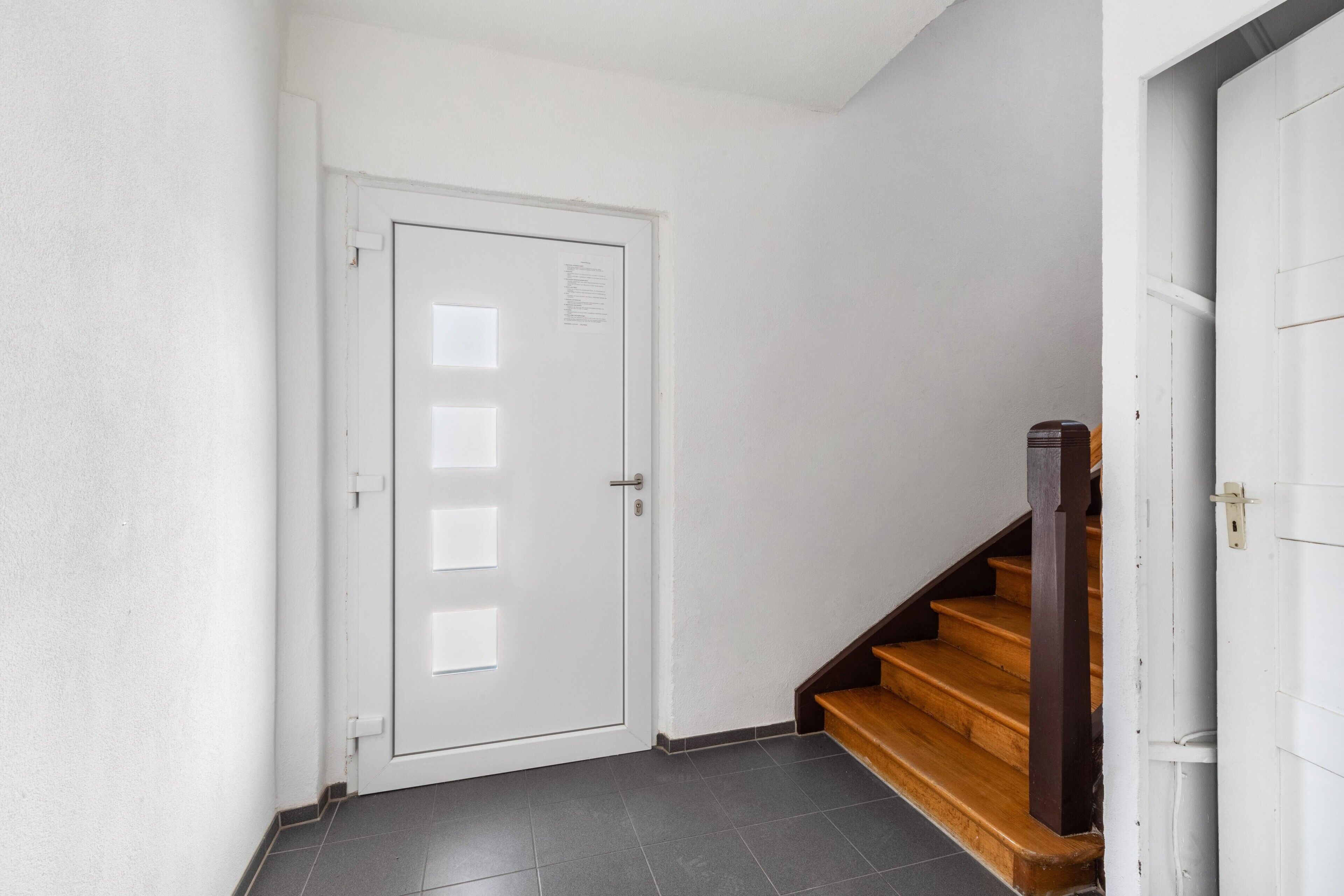 Una puerta blanca con un ventanal pequeño, una escalera de madera y un suelo de baldosas.