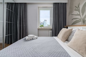 Ein modernes Hotelzimmer mit einem großen Bett, grauen Vorhängen, einem Fenster mit Blick auf die Stadt und einem kleinen Topfpflanzen auf dem Fensterbrett.