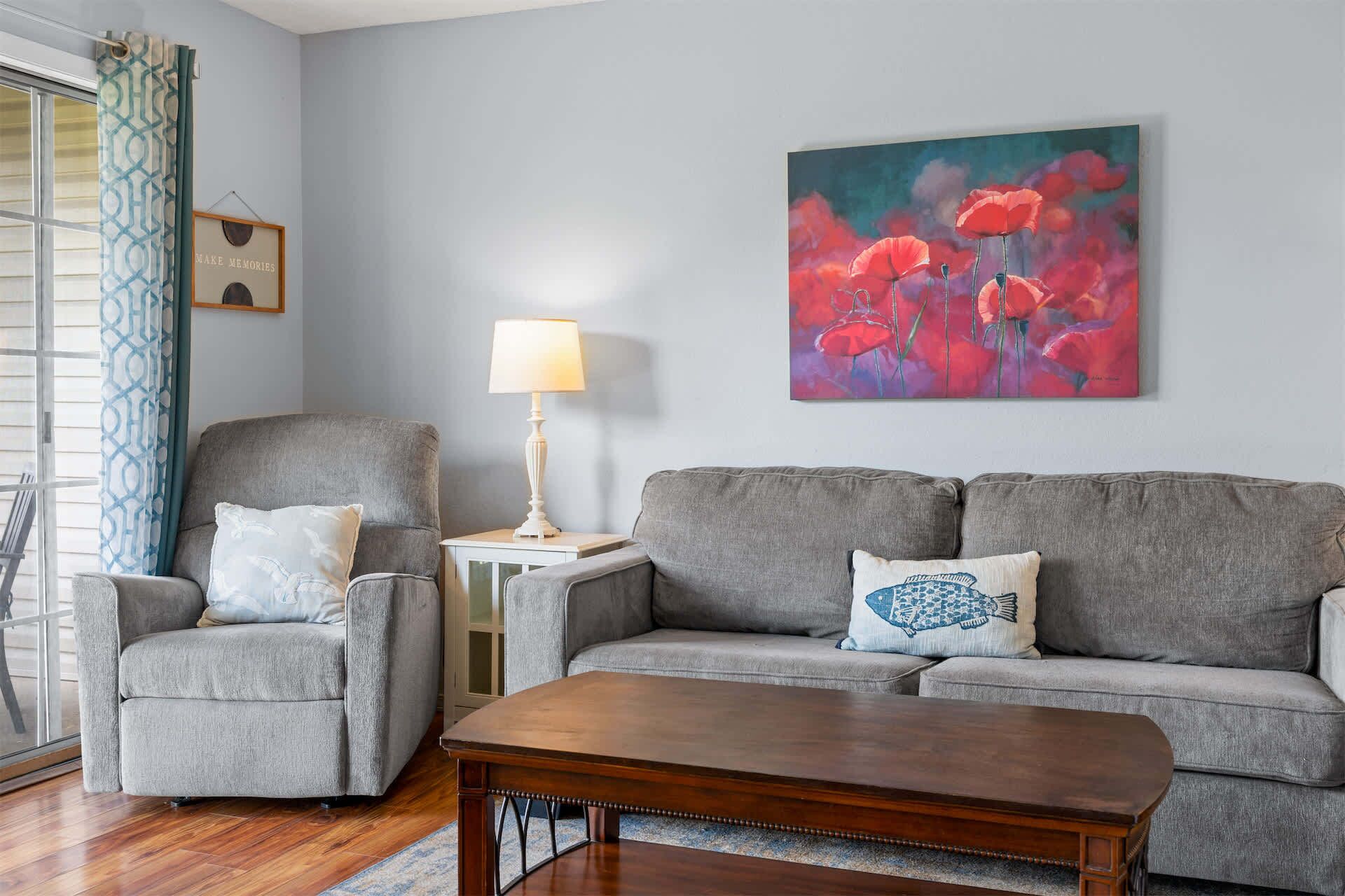Un salon avec un canapé gris, un fauteuil assorti, une table basse en bois et un tableau représentant des fleurs rouges accroché au mur.