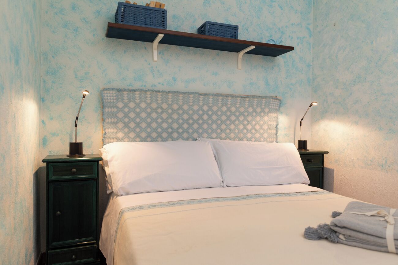 Un lit bien fait, avec du linge de lit blanc et une tête de lit à motifs, flanqué de deux tables de chevet avec des lampes, le tout adossé à un mur à texture.