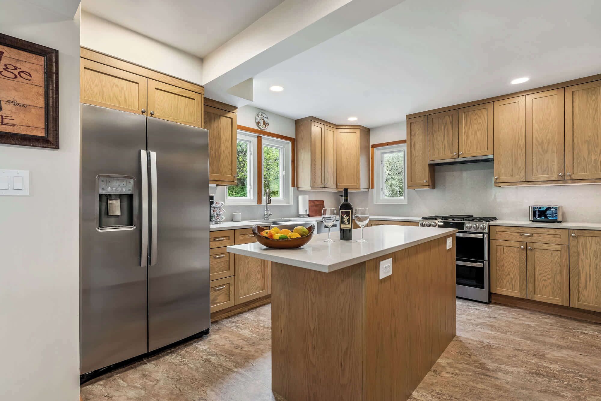 A modern kitchen with wooden cabinets, a stainless steel refrigerator, a built-in water dispenser, a large island with a bowl of fruit, and a wine bottle.