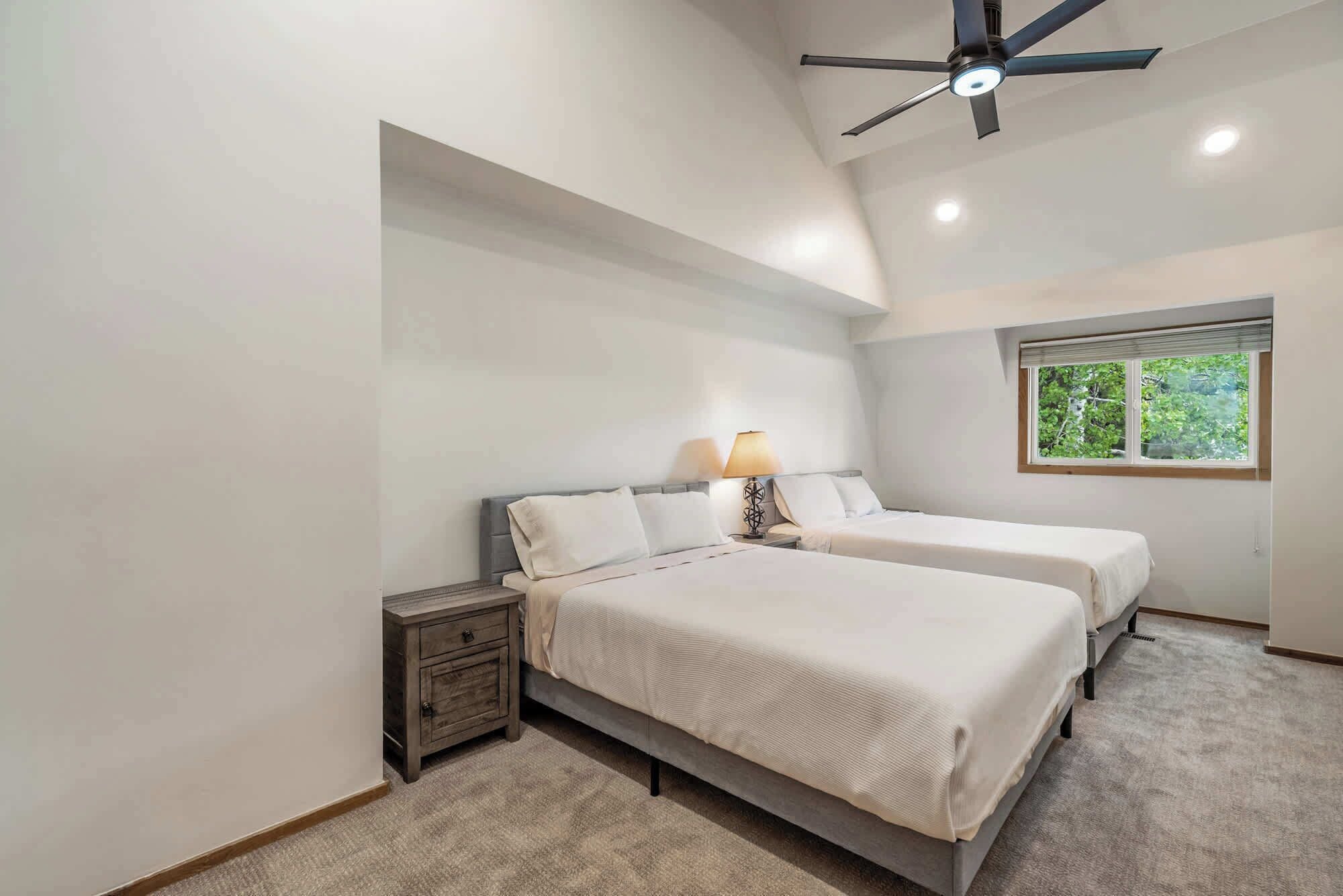A bedroom with two beds, a nightstand, a ceiling fan, a window with a view of greenery, and a lamp.