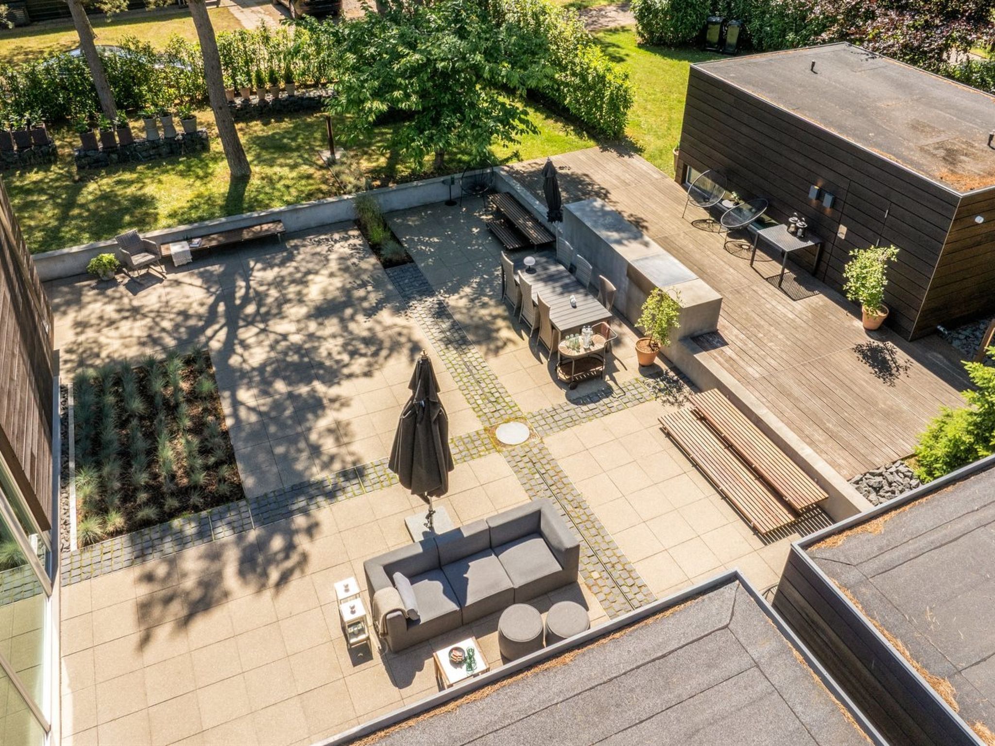 An outdoor patio with a sofa, tables, and chairs, surrounded by a garden and a building.