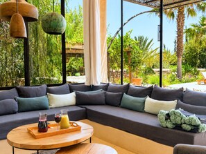 A modern living room with a sectional sofa, a round wooden coffee table, and a large window with a view of greenery and a pergola. - Palais Idalya (Essaouira)