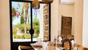 Zona de dinar amb taula de fusta per a quatre, vista a un jardí a través d'una finestra gran i una peça decorativa tradicional a la paret.