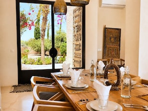 A dining area with a wooden table set for four, a view of a garden through a large window, and a traditional decorative wall piece. - Palais Idalya (Essaouira)