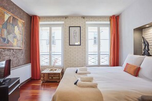 Ein Hotelzimmer mit einem großen Bett, einem Fernseher, einem Schreibtisch mit Lampe und Blick auf die Stadt durch große Fenster.