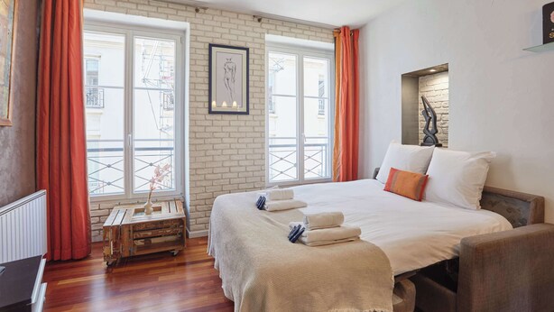 Ein Hotelzimmer mit einem großen Bett, einem Holfnachtkästchen, einem Heizkörper und Blick durch ein mit Vorhängen drapiertes Fenster.