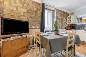 A dining area with a stone wall, a TV on a wooden dresser, a table set for two, and a kitchen in the background. - Apartment 'Centre Crozon' with Wi-Fi (Crozon)