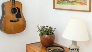 Una chitarra appesa al muro, un quadro incorniciato, un mobiletto in legno con cassetti, una pianta in vaso e una lampada da tavolo.