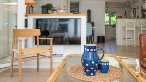 Un soggiorno moderno con una sedia in legno, un tavolino da caffè con un sottopiatti intrecciato e una brocca blu con pois bianchi.