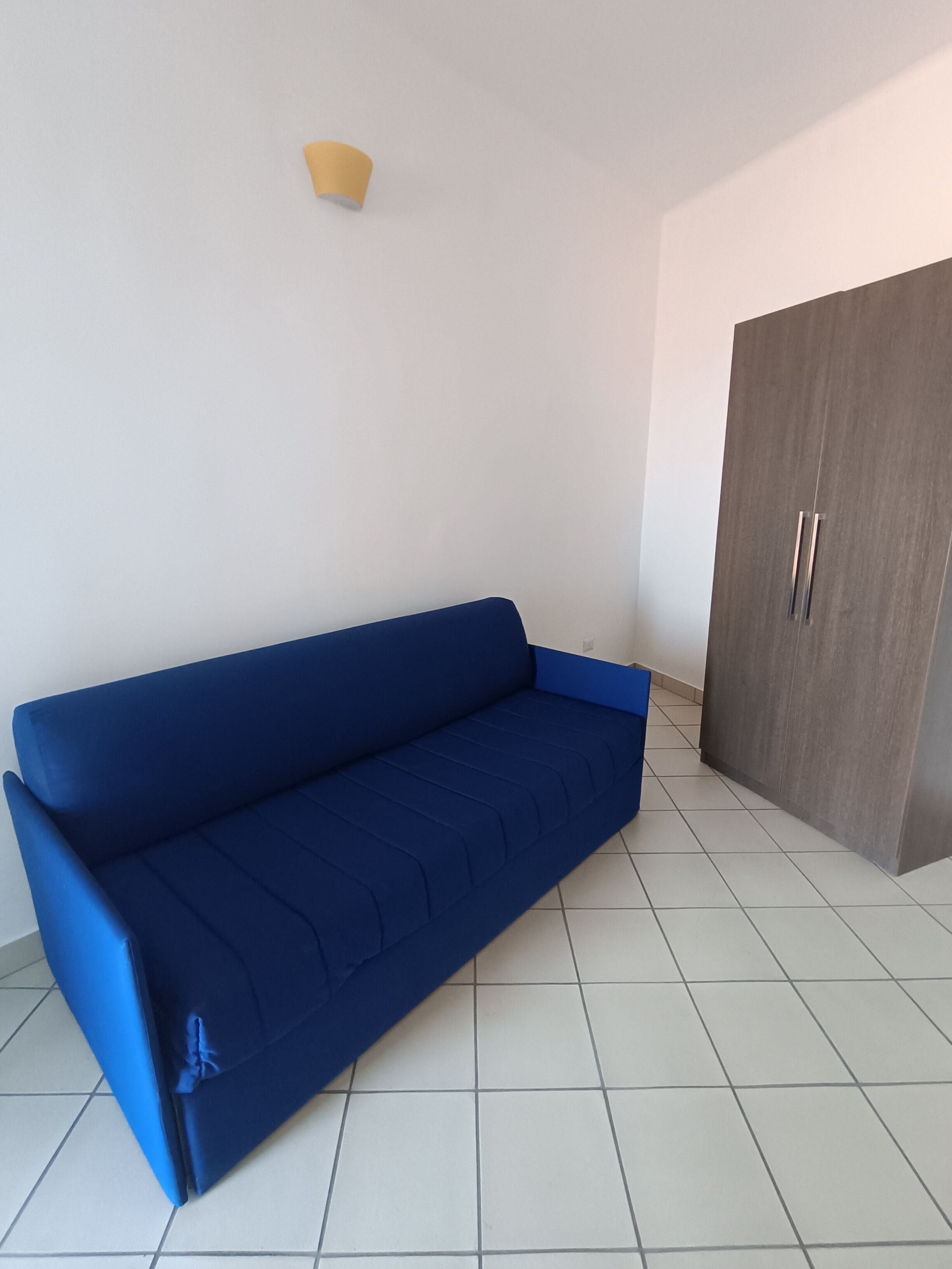 A blue sofa in a room with white tiled flooring and a wooden wardrobe.