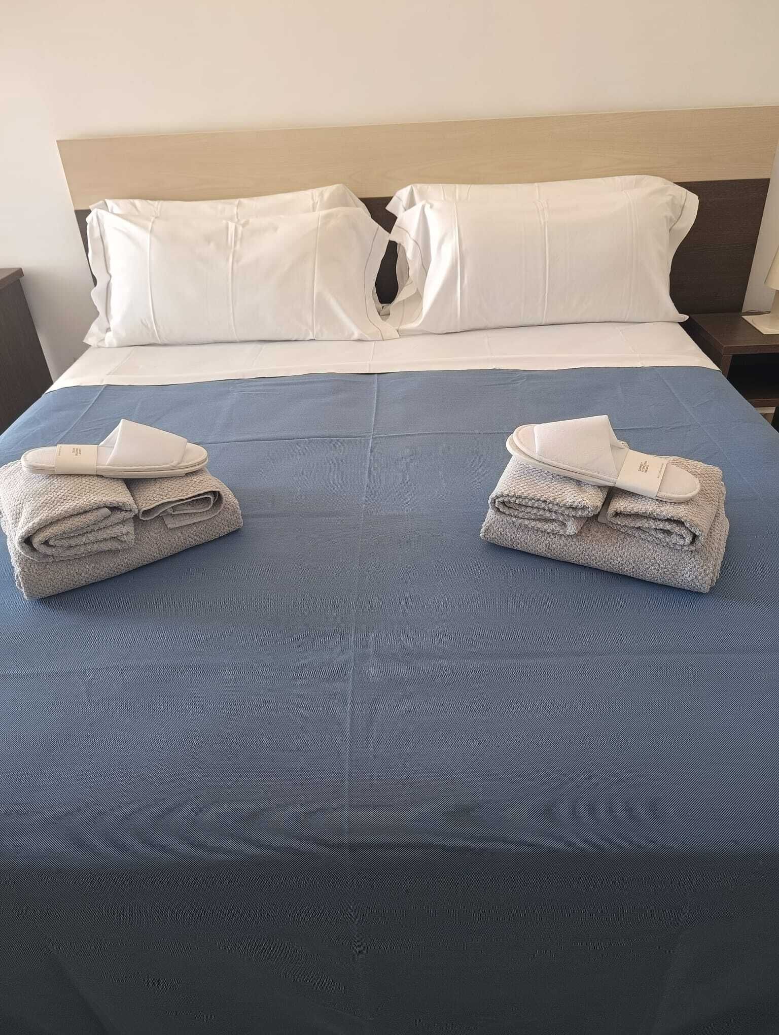 Un lit bien fait, recouvert d’une couverture bleue, avec deux serviettes pliées et une paire de chaussons de chaque côté.