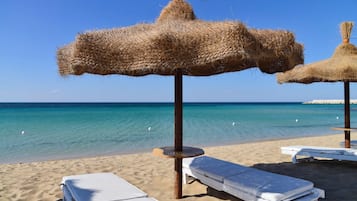 Una spiaggia con acque cristalline, sedie a sdraio bianche e ombrelloni di paglia.