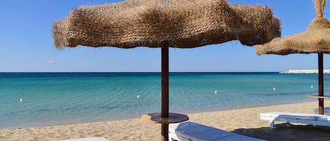 Ein Strand mit klarem, blauem Wasser, weißen Liegestühlen und Strohsonnenschirmen.