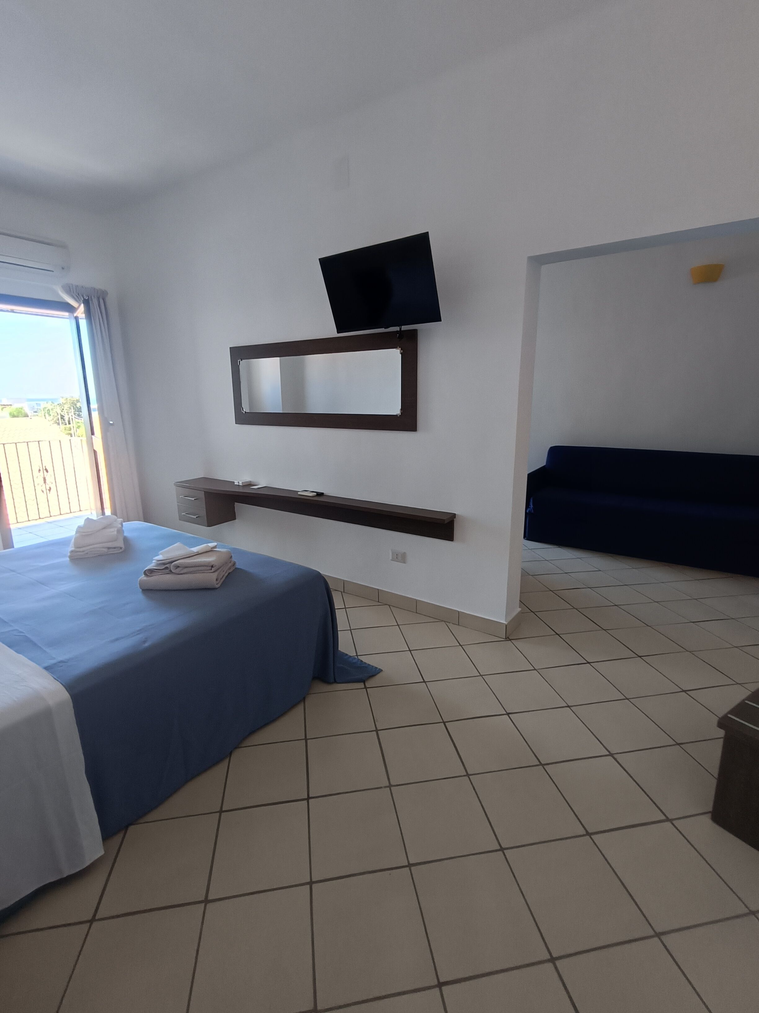 A hotel room with a bed, a TV mounted on the wall, a small shelf, and a view of the outdoors.