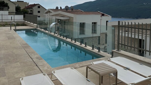 Una piscina con sillas de descanso, barandilla de cristal y vistas a casas y una colina.