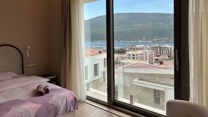 Habitación de hotel con cama, mesita de noche, ventana con cortinas y vistas a la costa con edificios y colinas.
