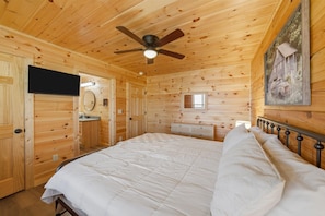 Une chambre dans une cabane en bois, avec un lit, un ventilateur de plafond, une télévision, une petite fenêtre et un tableau accroché au mur.