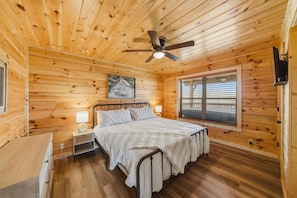 A wooden cabin bedroom with a bed, nightstands, a ceiling fan, a window with blinds, and a painting on the wall. - Morning Glory (Sevierville)