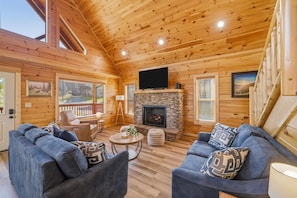 L’intérieur d’une cabane en rondins, avec une cheminée en pierre, un téléviseur à écran plat et un coin salon confortable.