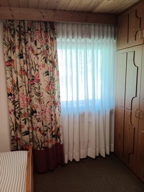 Ein Zimmer mit einem Bett, einem Holzschrank, einem Fenster mit durchscheinenden Vorhängen und einem blumigen Vorhang.