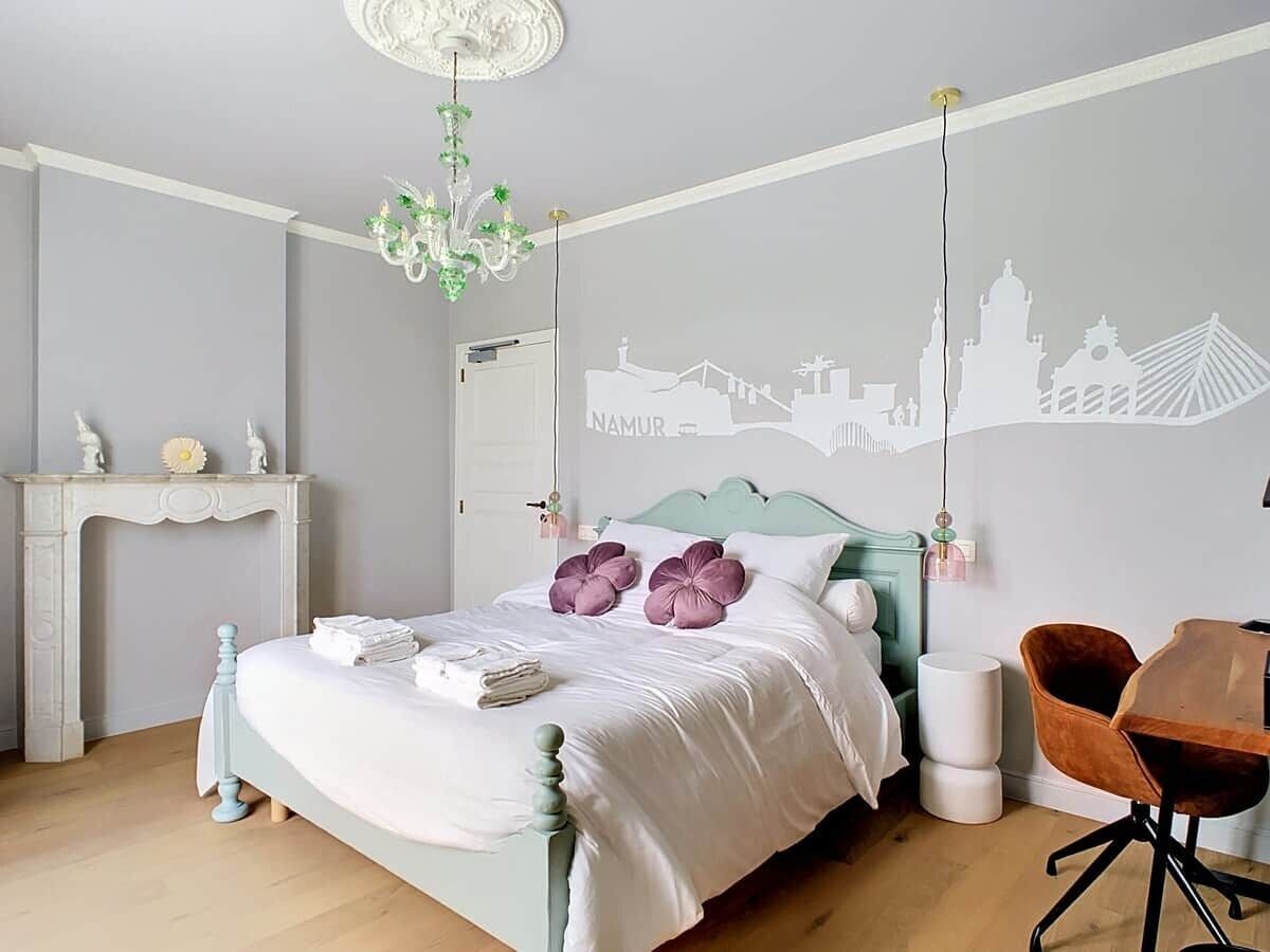 Une chambre à coucher comprenant un lit, une chaise, une cheminée et un mur orné d’une fresque représentant un paysage urbain.