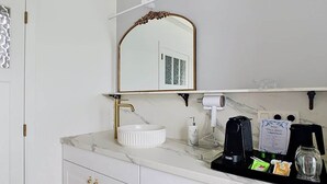 Ein Badezimmer mit einem weißen Marmorwaschbecken, goldenen Wasserhähnen, einem spiegel mit goldener Rahmung und einer schwarzen Kaffeemaschine auf der Ablagefläche.